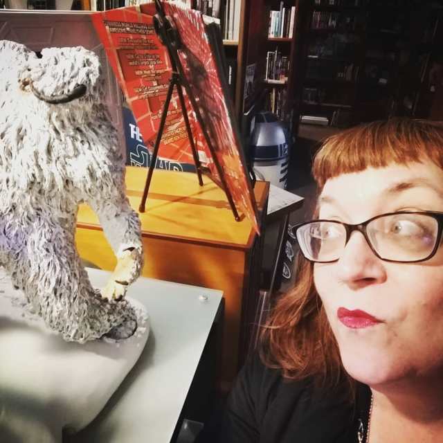 Pictured: Melissa Hillman, a redhead with short bangs, black-framed glasses, and red lipstick, gazes up and to the left at a small statue of a wampa, a creature from Star Wars. 